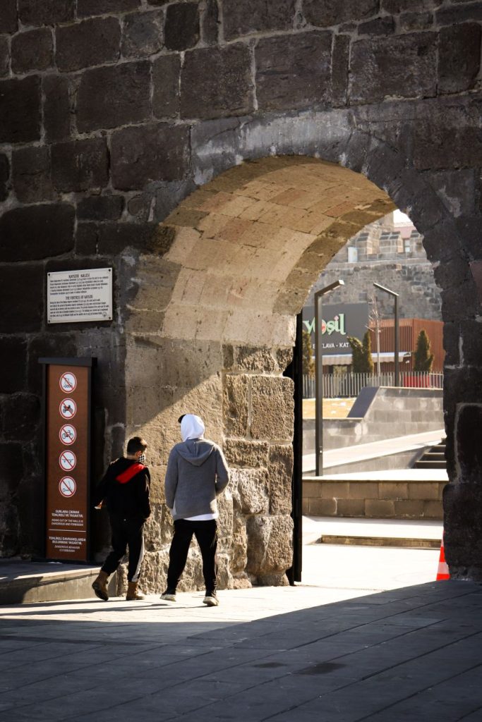 İki adam, güneşli bir günde taş bir kemer üzerinden rahat kıyafetlerle yürüyor.