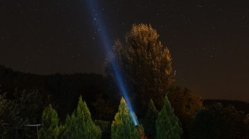 Bir kişinin yıldızlı gece gökyüzü altında, ağaçların çevresinde bir çadırın yanında fenerle kullandığı bir görüntü.