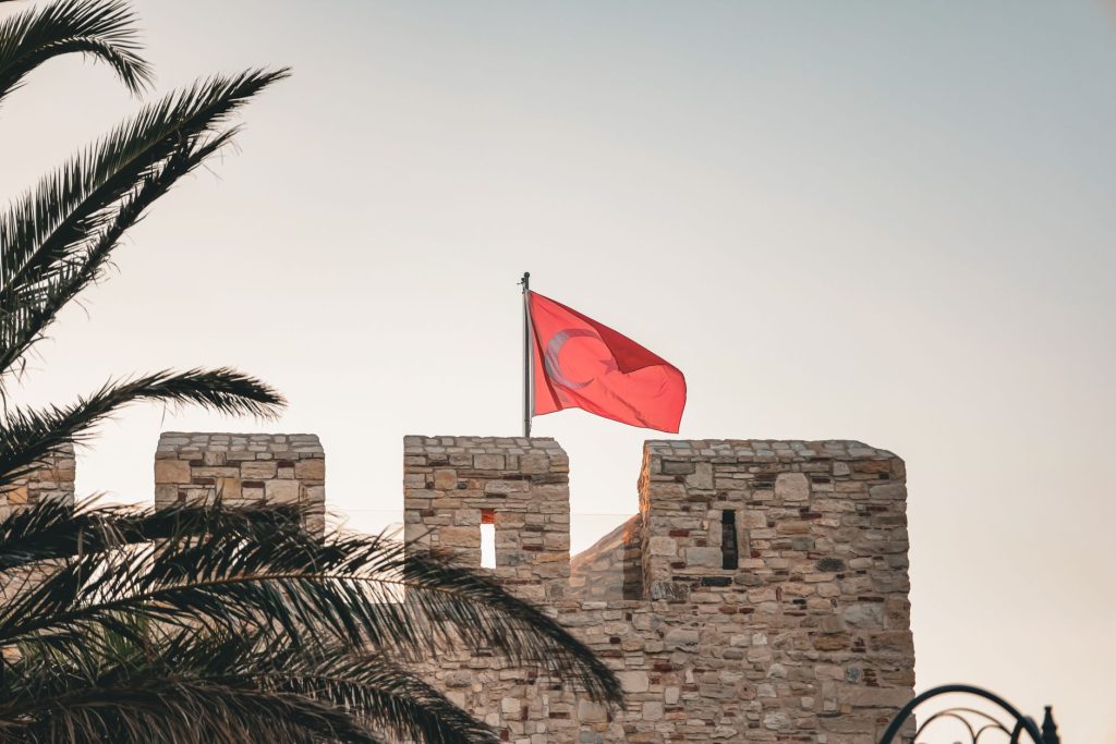 Palmiyelerin altında, berrak bir gökyüzünün altında tarihi bir taş kaledeki Türk bayrağı dalgalanıyor.