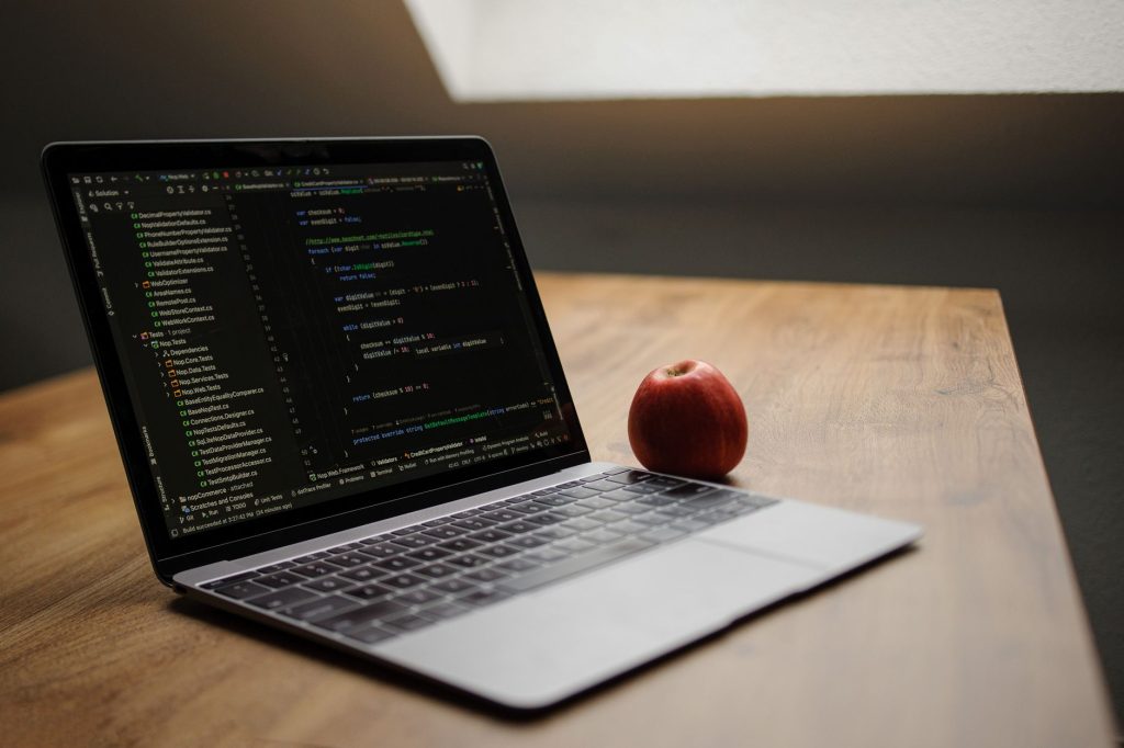 Açık laptopta kod gösterimi, ahşap masada kırmızı elmanın yanında.