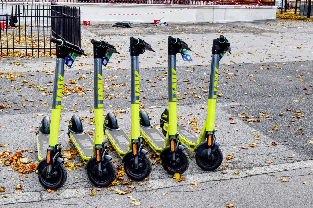 Sonbahar mevsiminde yapraklarla kaplı bir sokakta yan yana park edilmiş beş adet açık yeşil elektrikli scooter.