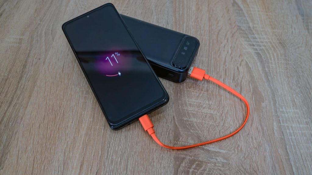 Ahşap bir masa üzerinde, turuncu bir kabloyla taşınabilir şarj cihazından şarj olan bir akıllı telefon.
