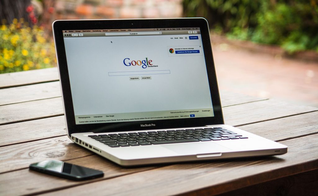 Ahşap bir masanın üzerine yerleştirilmiş bir MacBook Pro, dışarıda bir akıllı telefonun yanında Google Arama'yı gösteriyor.