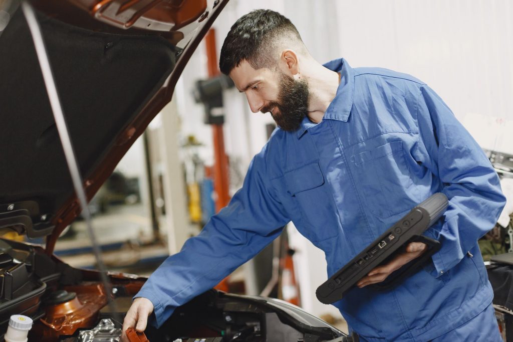 Mavi tulum giymiş bir oto tamircisi, bir garajda bir arabanın motorunu kontrol ediyor.