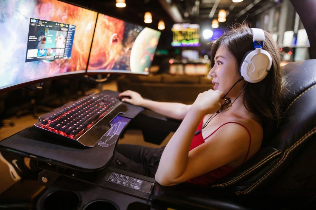 Kırmızı üst giymiş genç kadın, modern bir setup'ta beyaz kulaklıklarla çift monitörde oyun oynuyor.