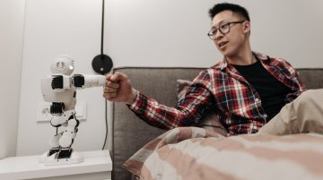 Asyalı bir adam modern bir yatak odasında robotla tokalaşarak yatakta oturuyor.