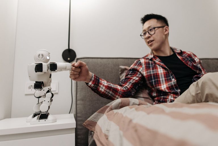 Asyalı bir adam modern bir yatak odasında robotla tokalaşarak yatakta oturuyor.